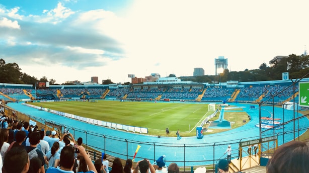 A large stadium with a well-maintained grass football field is surrounded by stands filled with spectators. The seating area is mostly blue, contrasting with yellow accents. In the background, urban buildings and trees are visible under a partly cloudy sky. People can be seen standing near the fence, some wearing sports jerseys.
