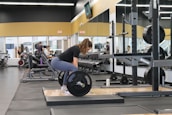 A woman is performing a deadlift exercise with a barbell in a gym. The gym is well-equipped, featuring various machines, weights, and mirrors lining the walls. Several other individuals are seen exercising in the background, contributing to a lively and focused atmosphere.