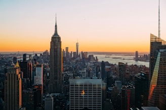 A panoramic cityscape of New York City at sunset, with skyscrapers silhouetted against a vibrant orange sky. The Empire State Building stands prominently in the foreground, and the horizon features distant urban structures along with a body of water.