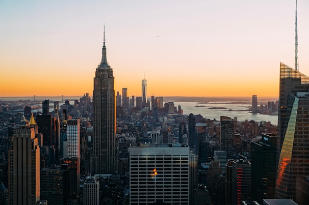 A panoramic cityscape of New York City at sunset, with skyscrapers silhouetted against a vibrant orange sky. The Empire State Building stands prominently in the foreground, and the horizon features distant urban structures along with a body of water.
