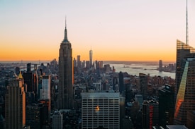 A panoramic cityscape of New York City at sunset, with skyscrapers silhouetted against a vibrant orange sky. The Empire State Building stands prominently in the foreground, and the horizon features distant urban structures along with a body of water.