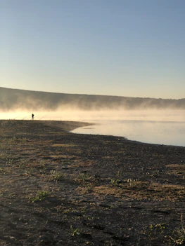 A rugged outdoor scene with a hunter and angler exchanging gear beside a misty forest river at dawn.