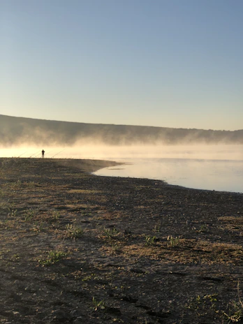 A serene early morning fishing scene by a misty lake with a lone fisherman casting a line.