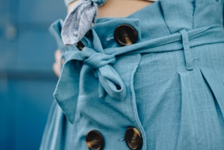 Close-up of a blue fabric with a tied belt and large button details. The material appears textured and has a loose, casual style.