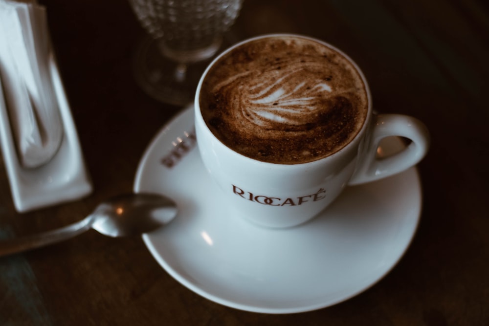 selective-focus photography of latte in white Riocafe cup on top of saucer near teaspoon