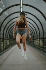 A person wearing a black tank top, denim shorts, and white sneakers with striped socks appears to be levitating within a futuristic, enclosed walkway. They are wearing a black cap, and the background comprises curved architectural elements.