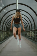 A person wearing a black tank top, denim shorts, and white sneakers with striped socks appears to be levitating within a futuristic, enclosed walkway. They are wearing a black cap, and the background comprises curved architectural elements.