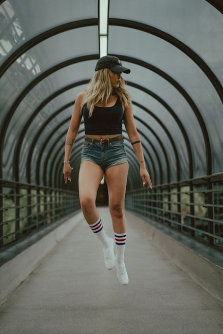 A person wearing a black tank top, denim shorts, and white sneakers with striped socks appears to be levitating within a futuristic, enclosed walkway. They are wearing a black cap, and the background comprises curved architectural elements.