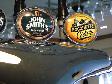 Two beer taps are prominently displayed, featuring labels for 'John Smith's Tadcaster Brewery' and 'Rocquette Cider' from the Channel Islands. The taps have a glossy metallic finish, reflecting light and the surrounding environment.