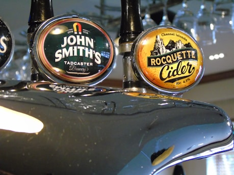 Two beer taps are prominently displayed, featuring labels for 'John Smith's Tadcaster Brewery' and 'Rocquette Cider' from the Channel Islands. The taps have a glossy metallic finish, reflecting light and the surrounding environment.