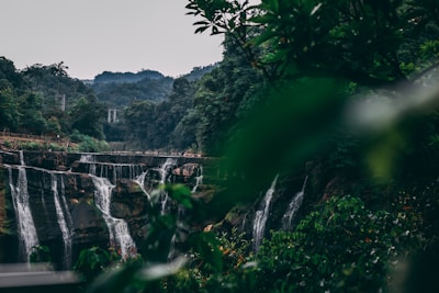 A lush green landscape featuring a cascading waterfall surrounded by dense forest and vegetation. The water flows over multiple rocky ledges. In the background, a suspension bridge can be seen crossing the valley, and hills create a serene backdrop.