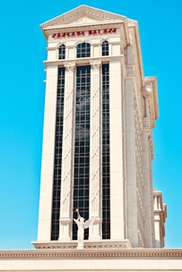 A tall, elegant building features classical architectural elements like columns and arches, with a prominent statue of an angel holding a trumpet in the foreground. The structure has a modern glass facade contrasted by traditional white stonework, and the words 'Caesars Palace' are visible at the top of the building. The sky is clear and bright blue, enhancing the building's grandeur.