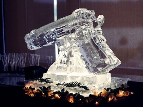 An intricately carved ice sculpture resembling a revolver is displayed on a decorative base. The sculpture is illuminated, contrasting its clear texture against a dark backdrop. Soft glowing lights and green foliage encircle the base, adding an elegant touch. Empty champagne flutes are visible in the background.
