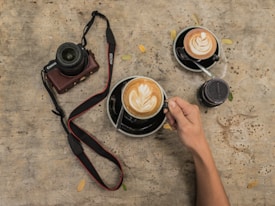 A rustic table surface is adorned with two cups of cappuccino, each showcasing intricate latte art. A hand is holding one of the cups. Nearby, a camera with a strap and a lens cap are placed, hinting at a photography theme. Scattered small leaves add to the earthy tone.