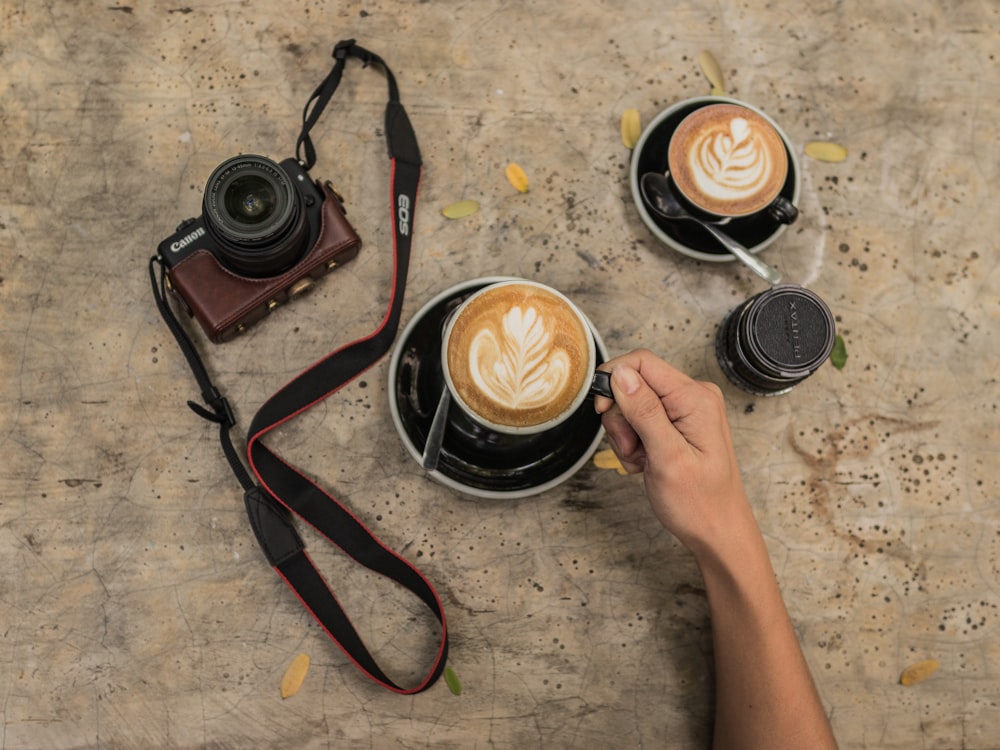 person holding black ceramic mug filled with latte on round black and white ceramic saucer near black and brown Canon DSLR camera