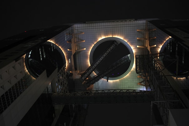 A modern architectural structure features geometric shapes with two large illuminated circular openings. The building appears to be part of an urban environment and has metal and glass elements. The lighting creates a dramatic effect against the nighttime sky.
