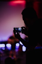 A silhouette of a person holding a smartphone, capturing a scene with a bright, blurred background. The phone screen displays a clearer image of what is being photographed, likely a performance or event with visible light sources.