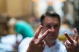 A person is in the background with their face blurred and their hands in focus, showing V signs with both hands. The setting appears to be in a busy, softly lit area, possibly a street or indoor market.