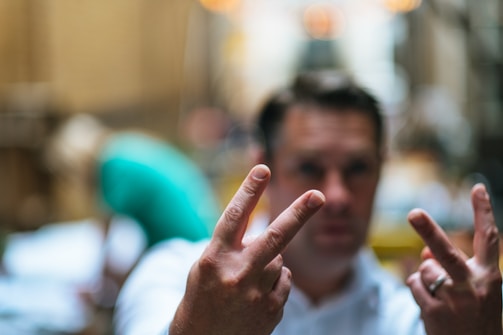 A person is in the background with their face blurred and their hands in focus, showing V signs with both hands. The setting appears to be in a busy, softly lit area, possibly a street or indoor market.