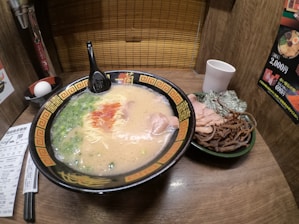 A bowl of ramen in a richly designed black and gold patterned bowl is placed on a wooden table. The ramen consists of a creamy broth with noodles, sliced pork, green onions, and drizzles of red sauce. A black spoon rests on the bowl. Next to the bowl is a side dish containing sliced pork, seaweed, and bamboo shoots. A white cup and a whole egg are also visible. The setting appears to be a cozy, individual dining booth with wooden walls and a bamboo screen.