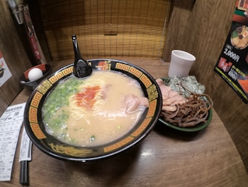 A bowl of ramen in a richly designed black and gold patterned bowl is placed on a wooden table. The ramen consists of a creamy broth with noodles, sliced pork, green onions, and drizzles of red sauce. A black spoon rests on the bowl. Next to the bowl is a side dish containing sliced pork, seaweed, and bamboo shoots. A white cup and a whole egg are also visible. The setting appears to be a cozy, individual dining booth with wooden walls and a bamboo screen.