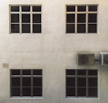 A beige wall with four dark-tinted windows arranged in two rows. Two air conditioning units are mounted on the right side. The surface of the wall has a textured appearance.