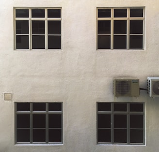 A beige wall with four dark-tinted windows arranged in two rows. Two air conditioning units are mounted on the right side. The surface of the wall has a textured appearance.