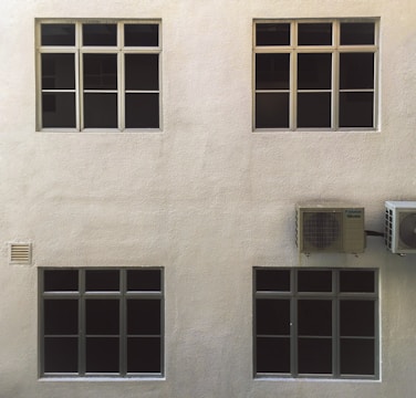 A beige wall with four dark-tinted windows arranged in two rows. Two air conditioning units are mounted on the right side. The surface of the wall has a textured appearance.