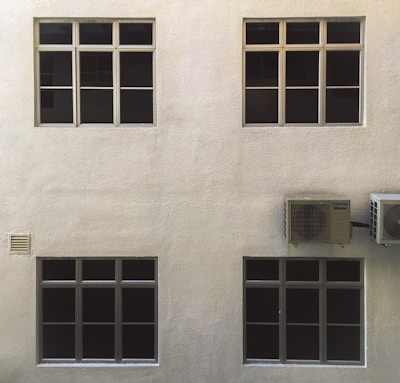 A beige wall with four dark-tinted windows arranged in two rows. Two air conditioning units are mounted on the right side. The surface of the wall has a textured appearance.
