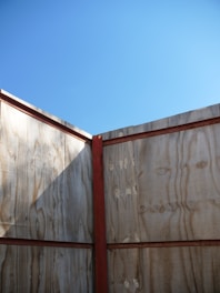 A wooden structure forms a corner with vertical planks and horizontal red beams. The light wood has visible grain patterns and a natural finish. Above, the sky is clear and blue, creating a stark contrast with the earthy tones of the wood.