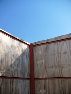 A wooden structure forms a corner with vertical planks and horizontal red beams. The light wood has visible grain patterns and a natural finish. Above, the sky is clear and blue, creating a stark contrast with the earthy tones of the wood.