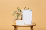 A wooden stool holds a variety of objects, including a decorative wooden reindeer figure, a piece of blank stationery adorned with a border and small leafy graphics, and a green leafy plant. The backdrop is a solid light brown color, creating a peaceful and minimalist scene.