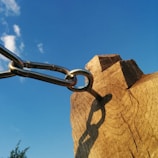 A chain connected to a wooden post against a clear blue sky. The metal chain links are silver, attached to a weathered piece of wood with visible grain and knots. The shadow of the chain is cast on the wood, creating a detailed silhouette.