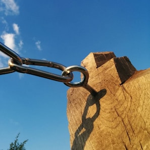 A chain connected to a wooden post against a clear blue sky. The metal chain links are silver, attached to a weathered piece of wood with visible grain and knots. The shadow of the chain is cast on the wood, creating a detailed silhouette.
