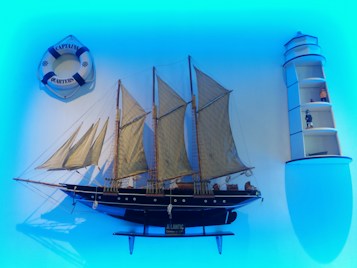 A decorative setup featuring a detailed model of a sailing ship with multiple masts and beige sails. To the left, there is a lifebuoy with text 'Captain's Quarters'. On the right, a lighthouse-shaped shelf displays small figurines and nautical objects. The scene is set against a blue backdrop, creating a maritime ambiance.