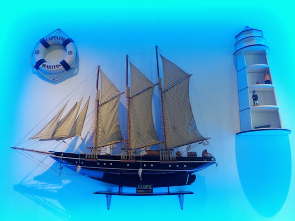 A decorative setup featuring a detailed model of a sailing ship with multiple masts and beige sails. To the left, there is a lifebuoy with text 'Captain's Quarters'. On the right, a lighthouse-shaped shelf displays small figurines and nautical objects. The scene is set against a blue backdrop, creating a maritime ambiance.