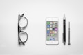 A minimalist arrangement of objects on a white background, including black-framed glasses, a smartphone displaying app icons, a black pen with metallic accents, and a small screwdriver.
