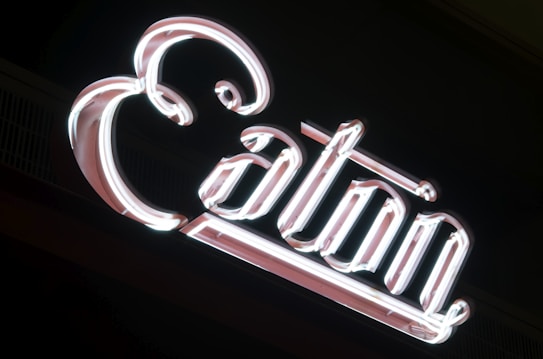 A neon sign with elegantly curved letters spelling out the word 'Col&oacute;n'. The sign emits a bright, white glow against a dark background.