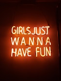 Neon sign displaying the phrase 'Girls Just Wanna Have Fun' in bright orange against a dark background.
