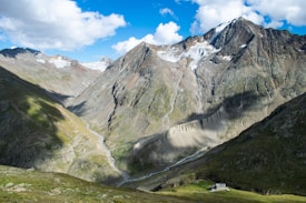 A stunning mountain landscape with steep, rugged peaks covered in patches of snow and ice. A deep valley with a flowing stream runs through the center, surrounded by lush green hills and rocky terrain. A small structure is visible nestled in the grass, adding a touch of human presence to the expansive natural scene. The sky is bright with scattered fluffy clouds, casting shadows over the mountains.