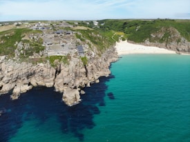 Rocky cliffs overlooking a serene turquoise sea with a small sandy beach nestled between rugged coastlines. There are buildings and a small road visible on the cliffs, surrounded by green vegetation. The area appears to be a quiet, picturesque coastal location.