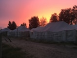 A series of large tents are set up on a dirt path with trees visible in the background. The sky is vividly colored with hues of orange and pink, suggesting either a sunset or sunrise. The tents are white and appear to be made of canvas. There are wires and poles indicating support structures, and shadows are cast due to the low light condition.