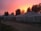 A series of large tents are set up on a dirt path with trees visible in the background. The sky is vividly colored with hues of orange and pink, suggesting either a sunset or sunrise. The tents are white and appear to be made of canvas. There are wires and poles indicating support structures, and shadows are cast due to the low light condition.
