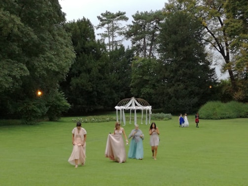 A group of people in formal attire walk across a green lawn. Among them, a few are wearing elegant dresses in pastel shades, while others in the background wear suits. Behind them stands a decorative white arch or canopy, possibly used for ceremonies. The area is surrounded by tall, lush green trees.