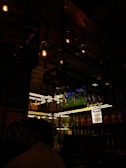 A dimly lit bar interior with bottles of liquor on shelves. Overhead, there are hanging light fixtures with exposed bulbs, casting a warm glow. Glasses are suspended above the bar, and a television screen shows a sports event faintly visible in the background.