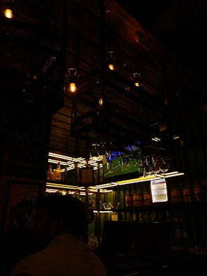 A dimly lit bar interior with bottles of liquor on shelves. Overhead, there are hanging light fixtures with exposed bulbs, casting a warm glow. Glasses are suspended above the bar, and a television screen shows a sports event faintly visible in the background.