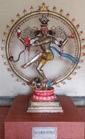 A detailed and colorful statue of a Hindu deity, adorned with intricate jewelry and clothing, surrounded by a circular frame with flames. The figure is posed in a dynamic dance, with multiple arms holding various objects. The statue is placed on a red pedestal with a plaque at the base.