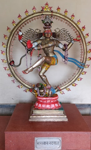 A detailed and colorful statue of a Hindu deity, adorned with intricate jewelry and clothing, surrounded by a circular frame with flames. The figure is posed in a dynamic dance, with multiple arms holding various objects. The statue is placed on a red pedestal with a plaque at the base.