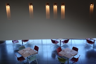 A modern dining space with four square tables arranged below. Each table is set with napkins and glassware, surrounded by chairs in various colors including red, green, and blue. The floor is a polished dark surface reflecting the faint blue light. The upper part of the image features a minimalist wall with five vertical, illuminated light fixtures evenly spaced.