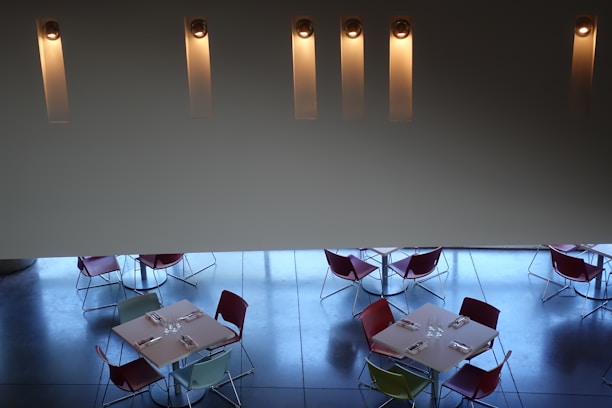 A modern dining space with four square tables arranged below. Each table is set with napkins and glassware, surrounded by chairs in various colors including red, green, and blue. The floor is a polished dark surface reflecting the faint blue light. The upper part of the image features a minimalist wall with five vertical, illuminated light fixtures evenly spaced.